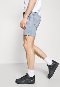 Shorts de mezclilla en un azul claro con dobladillos deshilachados, combinados con zapatillas negras y calcetines blancos. El diseño incluye bolsillos traseros y una silueta ajustada.