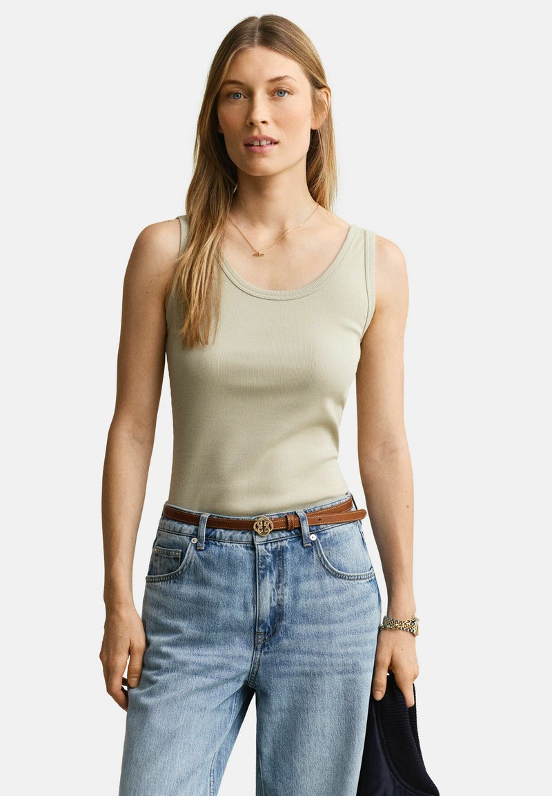 Femme aux longs cheveux blonds portant un haut beige sans manches, un jean bleu clair, une ceinture marron, tenant un vêtement foncé dans sa main gauche.