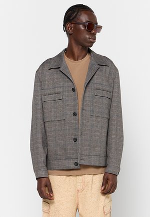 Uomo con capelli intrecciati che indossa occhiali da sole marroni, giacca grigia a quadri abbottonata, camicia color marrone chiaro e pantaloni cargo crema con fantasie.