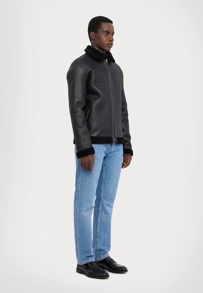 Veste en cuir noir avec un col en fourrure, fermeture éclair et poignets texturés, associée à un jean bleu clair et des chaussures noires.