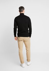 Muž v černém žebrovaném roláku, béžových kalhotách a bílých teniskách, stojící zády k jednobarevnému šedému pozadí.