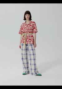 Femme debout portant une chemise à manches courtes rouge et blanche à motifs, un pantalon large à carreaux bleus, une ceinture jaune et des sandales vertes.