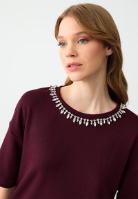 Bordeaux top met korte mouwen, met een met juwelen versierde halslijn afgewerkt met zilveren en heldere marquise-vormige versieringen.