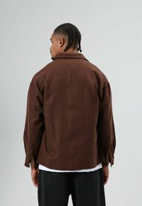 Bruine wollen overhemdjas met een losse pasvorm, kraag en met knopen afsluitbare manchetten. Draagt over een wit overhemd en gecombineerd met donkerblauwe jeans.