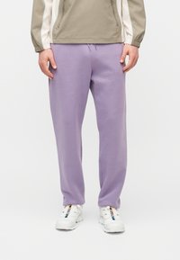 Άτομο που φοράει ανοιχτό μωβ φόρμες, λευκά αθλητικά παπούτσια με πολύχρωμες ρίγες και μπεζ-λευκό μπουφάν με φερμουάρ, όρθιο.