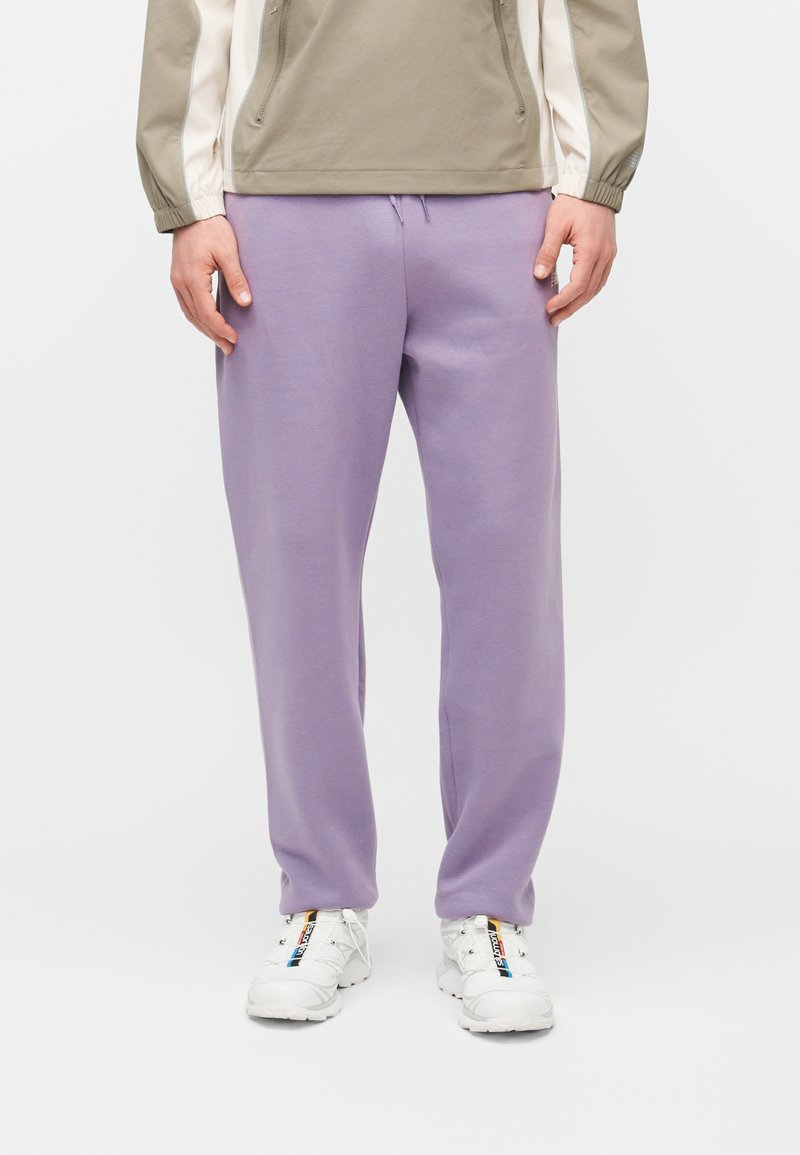 Άτομο που φοράει ανοιχτό μωβ φόρμες, λευκά αθλητικά παπούτσια με πολύχρωμες ρίγες και μπεζ-λευκό μπουφάν με φερμουάρ, όρθιο.