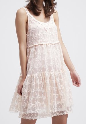 Kobieta ubrana w różową, bezrękawną sukienkę do kolan z kwiatową koronkową nakładką i falbanianym dołem, stojąca na jednolitym tle.