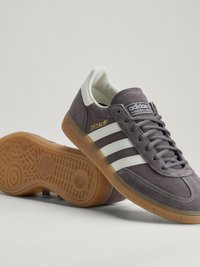Szara trampka Adidas Handball Spezial z białymi paskami, gumową podeszwą i sznurowadłami, jedna but stoi, a drugi leży na boku.