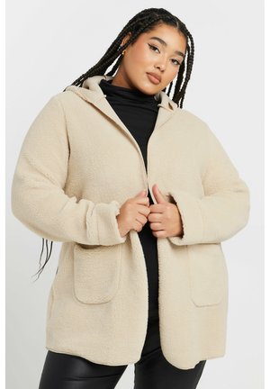 Veste en polaire beige avec capuche, coupe oversize, poches avant et texture douce. Le modèle la porte par-dessus un haut noir et un pantalon en cuir.