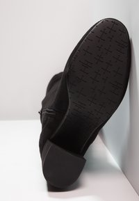 Bottine noire avec fermeture éclair latérale, motif de semelle visible, disposée en angle contre une surface et un mur blancs.