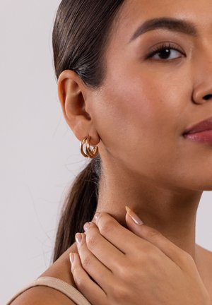 Gold-tone double hoop earrings on a model. Skin tone is warm, hair is dark and smooth, with hands gently touching the neck. Simple backdrop.