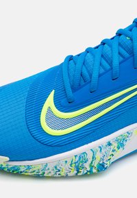 Blue athletic shoe with textured fabric, yellow accents, and mesh Nike swoosh. Colorful, patterned sole with white base.