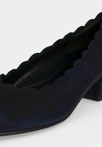 Chaussure en daim bleu marine à talon bas avec des bords festonnés et une semelle intérieure texturée, inclinée pour montrer les détails latéraux et intérieurs.