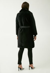 Cappotto in pelliccia sintetica nera con colletto, vita cintata, maniche lunghe e tessuto texturizzato. Indossato sopra leggings neri e scarpe con tacco alto.