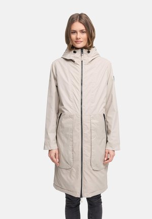 Donna che indossa un impermeabile lungo con cappuccio di colore beige chiaro, con cerniera frontale e due tasche con cerniera, in piedi davanti a uno sfondo neutro.