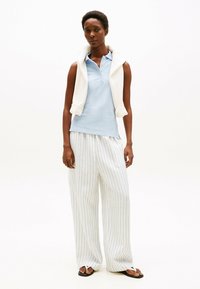 Polo bleu clair, pull blanc drapé sur les épaules et pantalon large à rayures. Des tongs noires complètent la tenue.