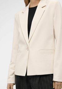 Blazer beige con una silueta estructurada, cierre de un solo botón y dos bolsillos frontales. Textura suave y solapas marcadas.