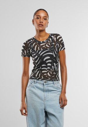 Femme portant un haut noir transparent avec des motifs abstraits de feuilles blanches et un jean taille haute bleu clair sur un fond gris uni.