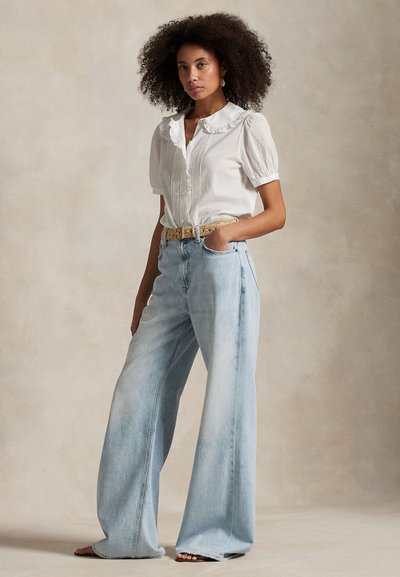 Kvinde med krøllet hår iført en hvid flæset bluse, lyseblå jeans med vide ben, beige flettet bælte og øreringe, som står med den ene hånd i lommen.