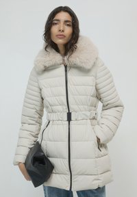 Cappotto invernale imbottito beige con colletto in pelliccia, vita cinte e dettagli neri con zip. Indossato con una pochette nera e jeans blu.