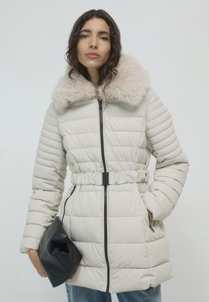 Manteau d'hiver beige rembourré avec un col en fourrure, taille ceinturée et détails zippés noirs. Porté avec une pochette noire et un jean bleu.
