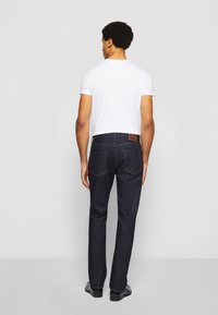 Dark blue denim jeans with a straight leg, five-pocket design, and a brown leather patch at the back waist, paired with a white t-shirt.