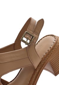 Sandalo in pelle tan con chiusura a fibbia, design con punta aperta e tacco in stile legno con texture. Cuciture visibili accentuano le giunture.