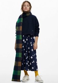 Haditengerészeti kék kábelszerű mintájú, rövidített pulóver, mintás sötétkék ruha fehér margarétákkal, zöld és barna kockás sál, sárga zoknik és fehér sportcipők.