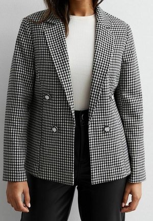 Kobieta ubrana w czarno-biały dwurzędowy blezer w jodełkę na białej prążkowanej bluzce i czarnych spodniach, stojąca na jednolitym tle.