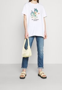 Camiseta blanca con un gráfico colorido de un ciervo y el texto "SOY UN CIERVA QUEER". Combinada con jeans azules, un bolso amarillo y sandalias amarillas con volantes.