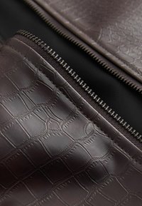 Bruin krokodillenleer met een gestructureerde afwerking, voorzien van een zwarte stofvoering en een zilveren rits langs de rand.