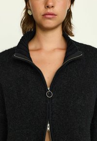 Schwarze Zip-Up-Strippe mit geripptem Kragen, aus strukturiertem Stoff gefertigt. Verfügt über einen silbernen Reißverschluss und runde metallische Akzentdetails.