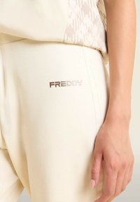 Joggers color beige claro hechos de un tejido suave, con un diseño elegante que presenta el logo "FREDDY" en metalizado plateado en la costura lateral.