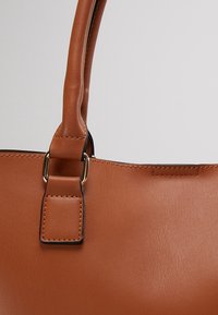 Sac à main en cuir marron avec une bandoulière cousue attachée par des anneaux métalliques dorés sur un fond gris clair uni.