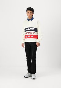 Krem genser med marineblå, rød og grå tekstbånd som leser "GANT" og "SPORT U.S.A." over svarte jeans og grå sneakers. Blå skjorte med krage under.