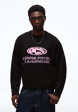 Junger Mann trägt schwarzen Pullover mit pinkem Text und Logo, dunkle Jeans, silberne Kettenhalskette und kleine Creolen vor weißem Hintergrund.