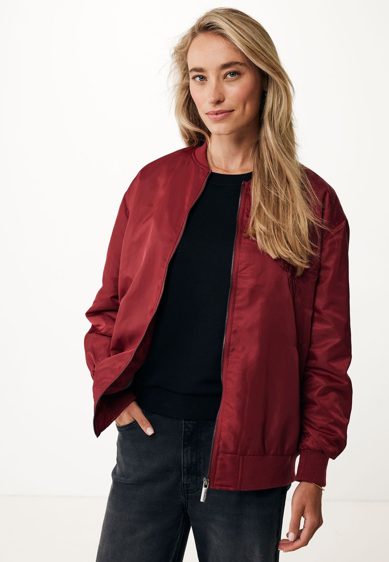 Veste bombardier bordeaux avec fermeture éclair à l'avant, poignets côtelés et texture lisse et brillante. Associée à un pull noir et un pantalon gris.