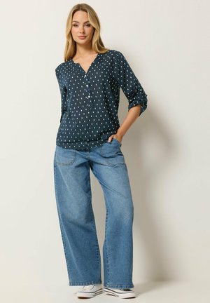 Mujer con cabello largo y rubio, vestida con una blusa azul marino con lunares, jeans azul de pierna ancha y zapatillas blancas, de pie con una mano en el bolsillo.