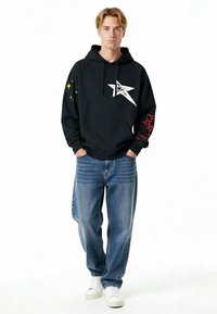 Sweat à capuche noir avec un graphisme d'étoile blanche et des accents d'étoiles rouges sur les manches, associé à un jean denim bleu clair et des baskets blanches.