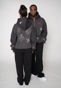 Grauer Kapuzenpullover mit Grafikdrucken in verschiedenen Farben, kombiniert mit schwarzen Jogginghosen. Verfügt über eine lockere Passform und dezente strukturierte Details.