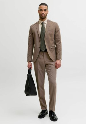 Homme en costume beige avec chemise beige, cravate à motifs, chaussures habillées noires, tenant un sac fourre-tout en cuir noir sur fond blanc.