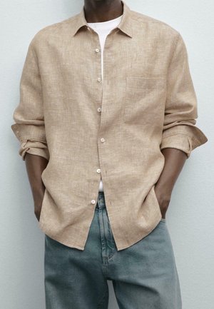 Mężczyzna w beżowej lnianej koszuli na guziki noszonej na białym t-shircie z podwiniętymi rękawami, w połączeniu z wyblakłymi niebieskimi dżinsami, stoi z rękami w kieszeniach.