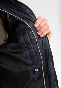 Marineblaue Innenfutter einer Jacke mit beigem Paspel, ausgestattet mit einer aufgesetzten Tasche, die in einer Hand gehalten wird und die glatte Textur und die Nähdetails zeigt.