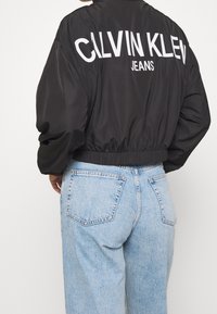 Černá saténová bomber bunda s bílým logem "CALVIN KLEIN JEANS" na zadní straně, s elastickým lemem a převislými rukávy, kombinovaná se světle modrými džínami.