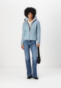 Lichtblauwe pufferjack met horizontale quilt, frontaal rits en een capuchon. Gecombineerd met lichtgewassen flared jeans en een zwarte tas.