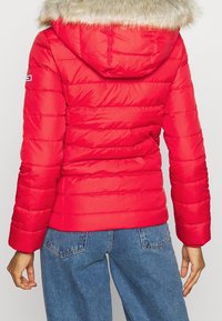 Chaqueta acolchada roja con capucha forrada de piel, con costuras horizontales y puños elásticos. Se lleva con vaqueros de mezclilla azules, vista trasera.