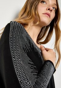 Giovane donna con capelli castano chiaro che indossa un maglione nero decorato con intricate fantasie di strass argentati sulle maniche e sul petto.