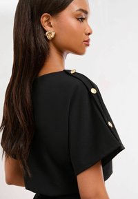 Crna majica s kratkim rukavima, glatka tkanina i dekorativne zlatne dugmadi na ramenu. Duga, ravna kosa vidljiva u profilu.