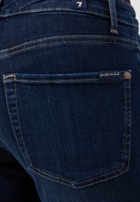 Jeanși din denim albastru închis, având un buzunar la spate cu un eticheta decorativă pe care scrie "pentru toți oamenii" și cusături subtile în contrast.