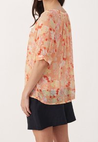 Blouse florale en pêche avec des motifs rouges et jaunes, manches courtes et coupe décontractée. Tissu léger avec une texture légèrement transparente.